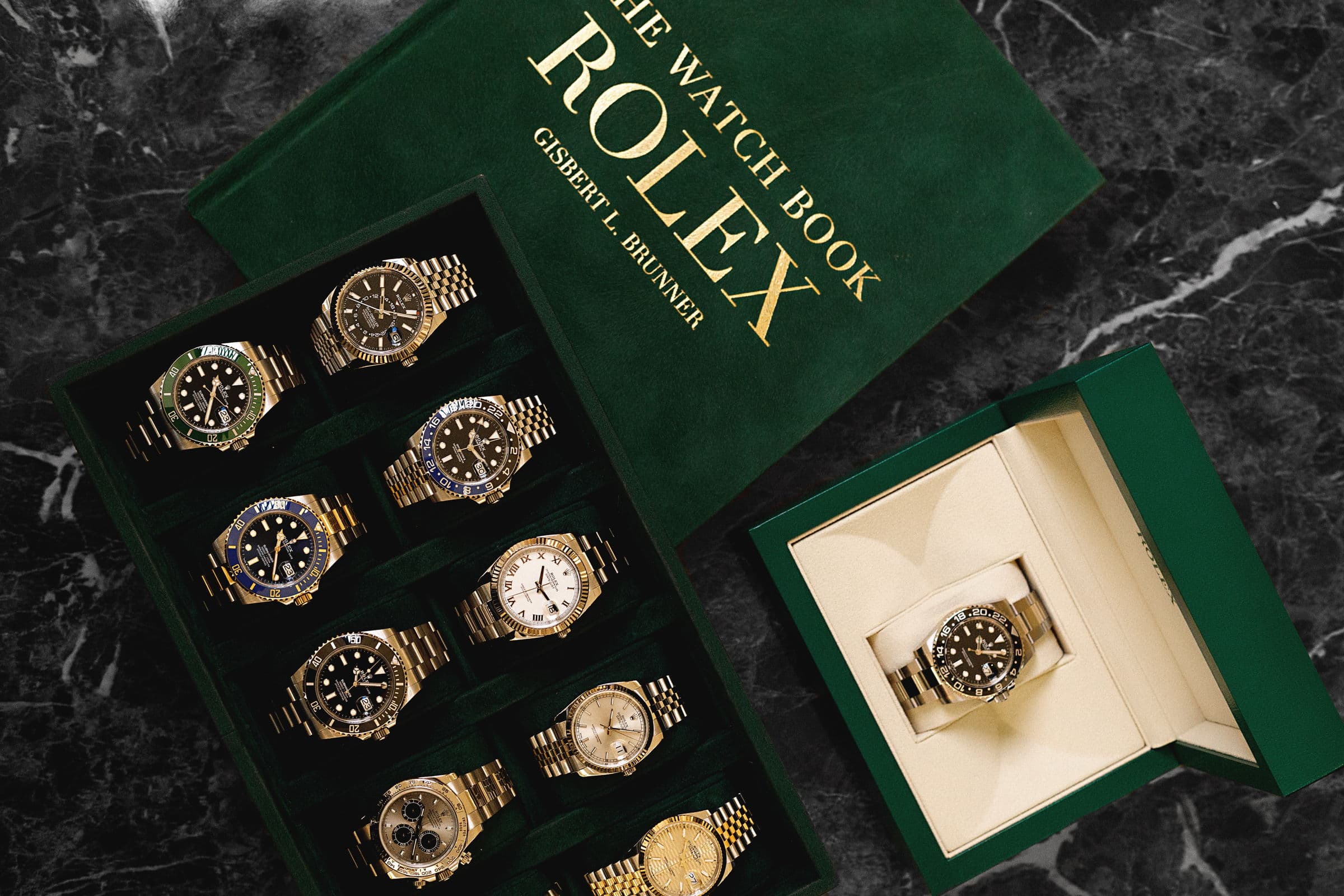 A tray of Rolex sports models beside The Watch Book, photographed on dark marble